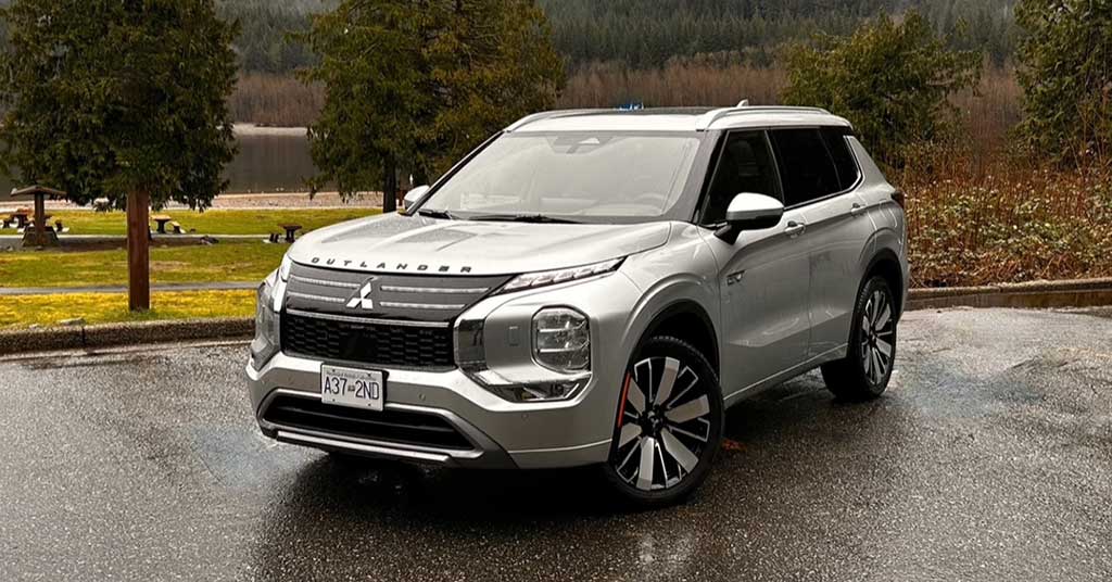 2026-Outlander-PHEV - Warman Voice Front-three-quarter view of a silver Mitsubishi Outlander SUV parked on a wet pavement with trees and a lake in the background, daytime outdoor scene.
