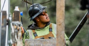 Construction worker in a hard hat, sunglasses, and green hoodie looks up at equipment on a construction site.